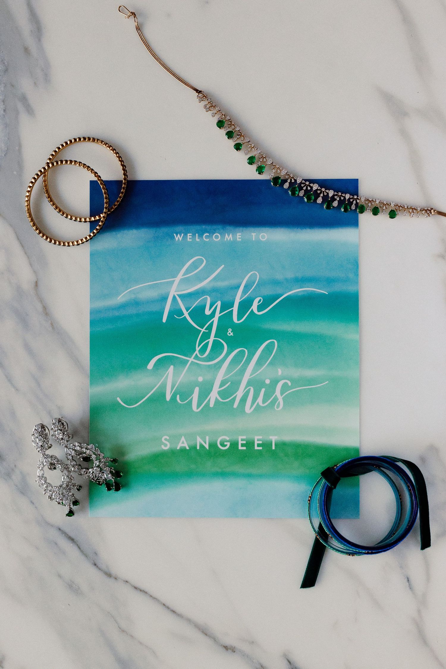 Watercolor wedding invitation for Kyle and Nikhis Sangeet surrounded by gold bangles and jewelry on marble.