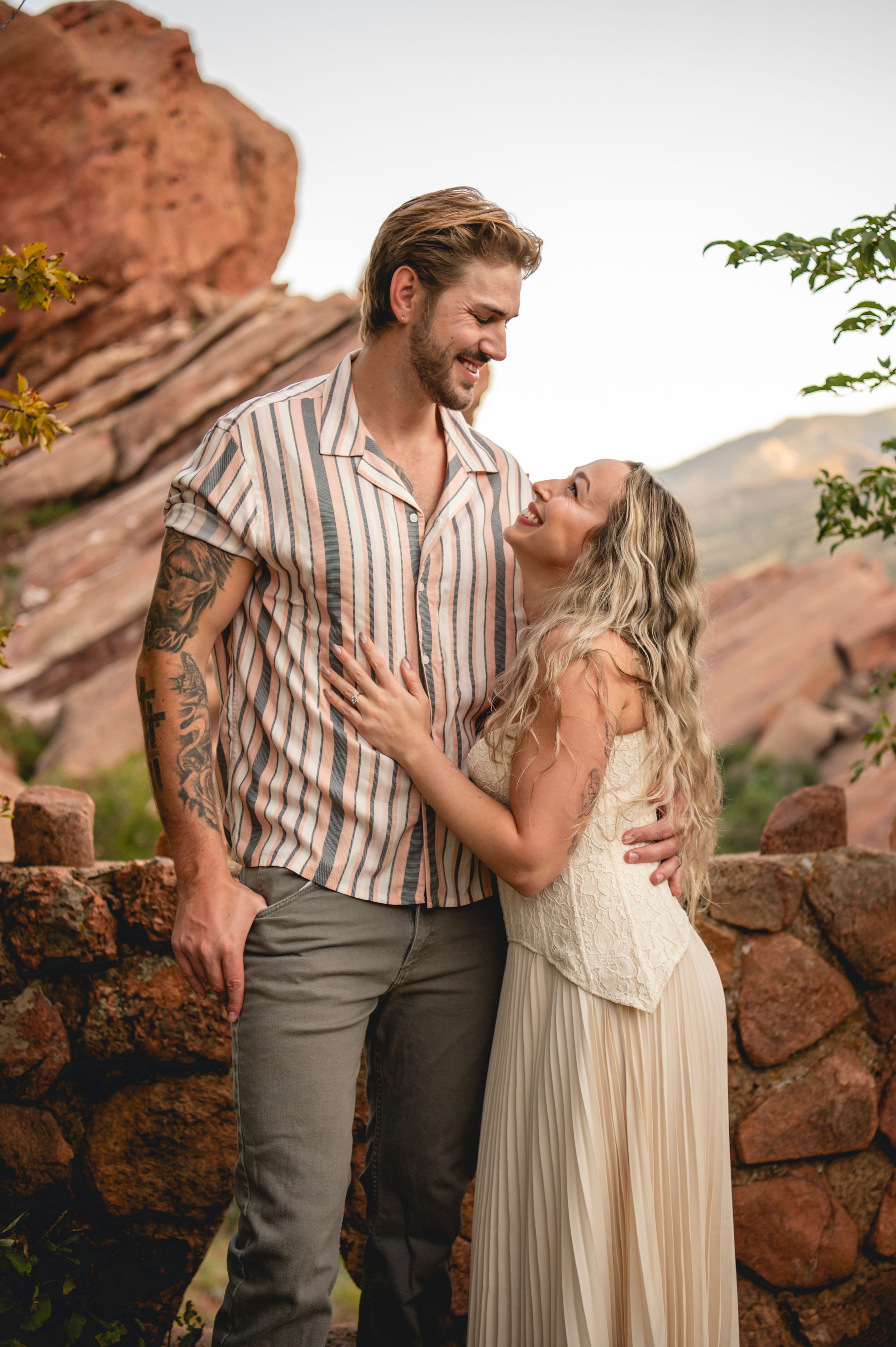 Couple embracing and laughing together at Red Rocks, man with tattoos in striped shirt, woman in white dress.