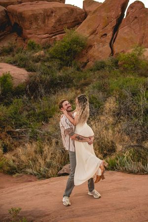 A couple dances joyfully among red rock formations, the woman in a flowing white dress being lifted by her tattooed partner.