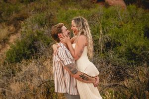 A couple laughs joyfully outdoors, the man lifting the pregnant woman in a white dress among desert shrubs.