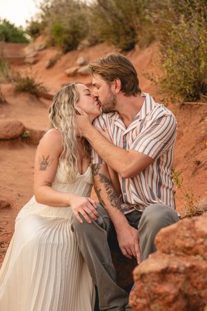 Couple kissing romantically on red rock desert landscape, woman in white dress with tattoos, man in striped shirt.