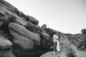 Couple embracing on a desert trail surrounded by large rock formations in a black and white landscape photo.