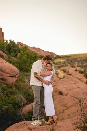 Couple embracing on red rock formation at Red Rocks Park, woman in white dress, golden hour sunset backdrop.