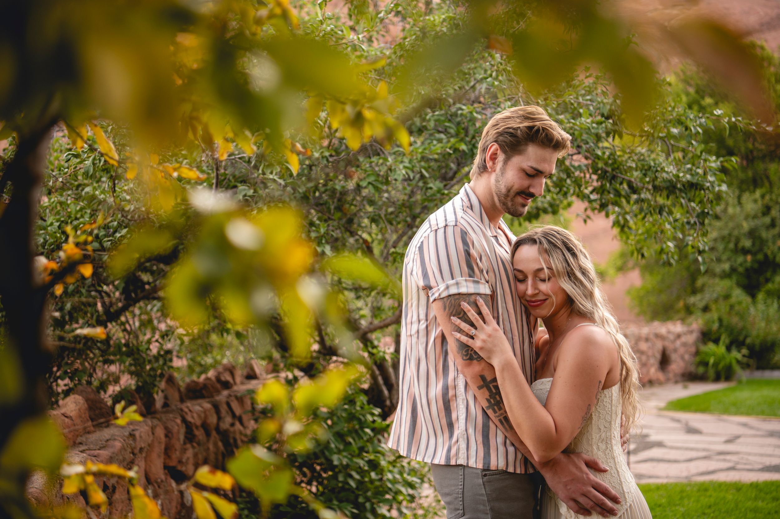 Couple sharing a romantic moment in a lush garden surrounded by colorful autumn foliage and red stone walls.