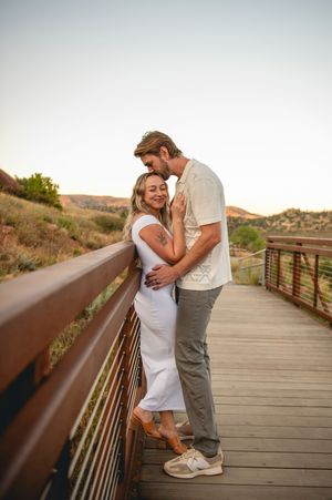 Couple embracing on a wooden bridge at sunset with red rock hills in the background.