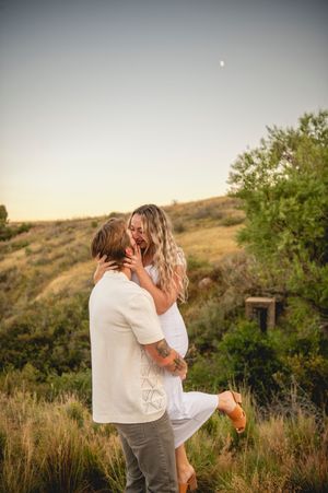 Couple embracing tenderly outdoors at golden hour with green hills and crescent moon.