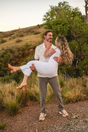 A man lifts a woman in white outdoors, both smiling, surrounded by lush greenery and golden grass at sunset.