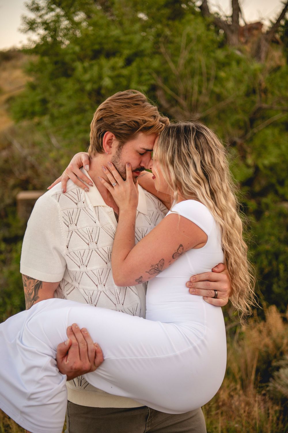 Romantic couple kissing closely while embracing in a lush outdoor setting at dusk.