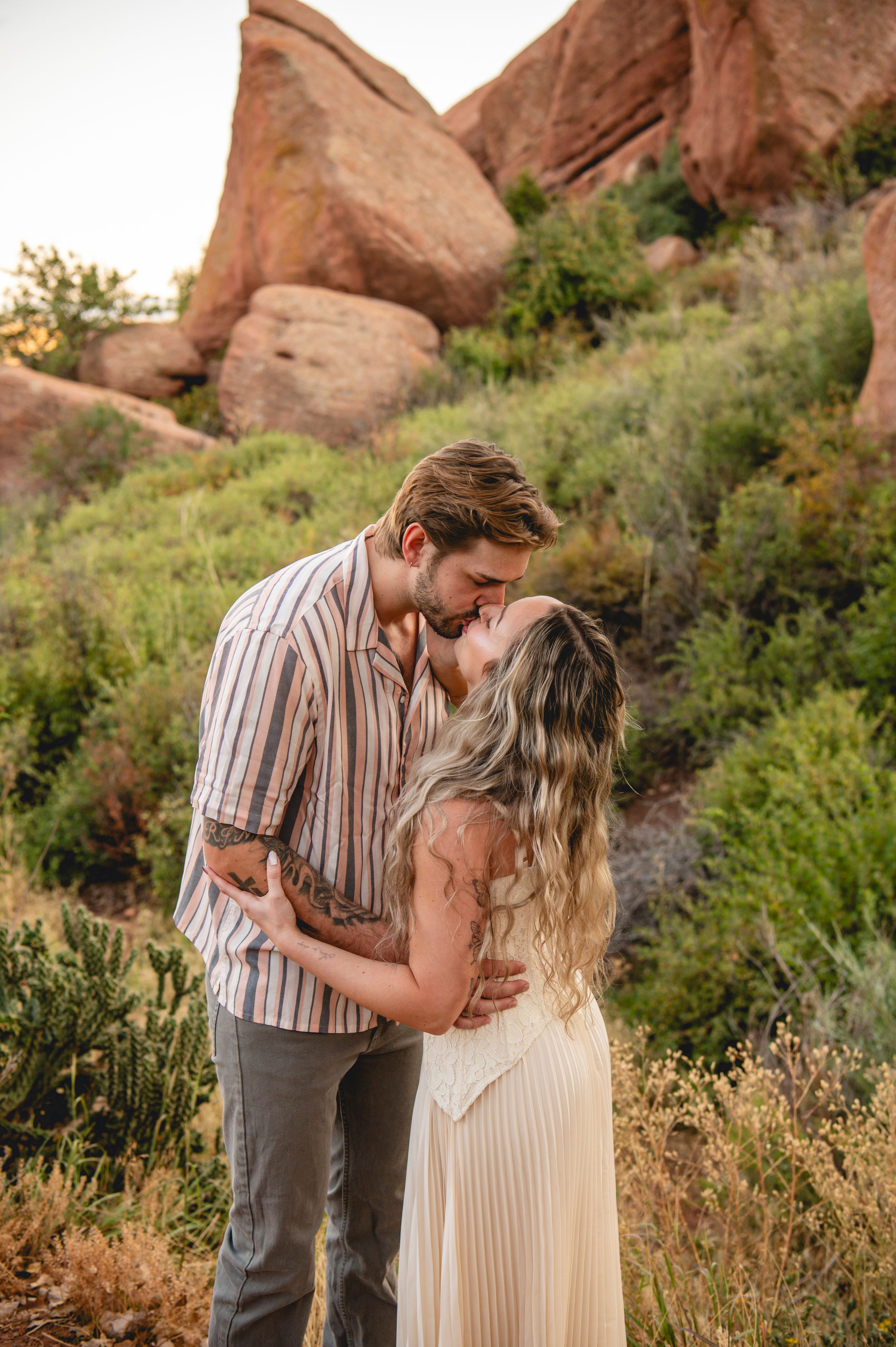Couple sharing a romantic kiss among red rock formations and lush greenery in a scenic outdoor engagement photo.