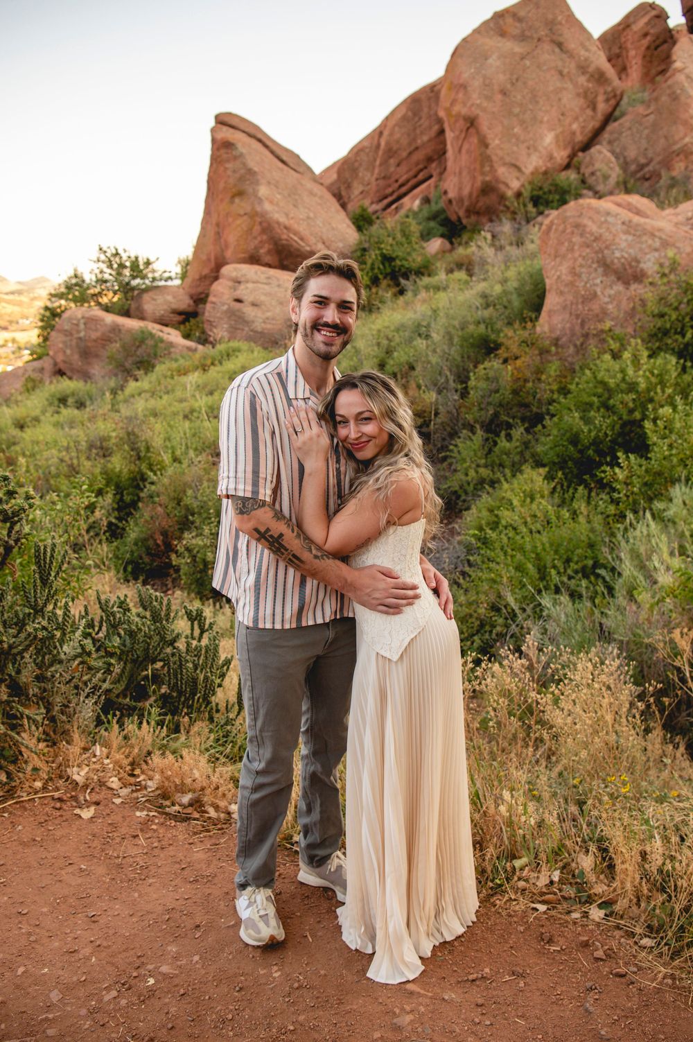 A couple embraces and smiles on a desert trail surrounded by red rocks and lush greenery at golden hour.