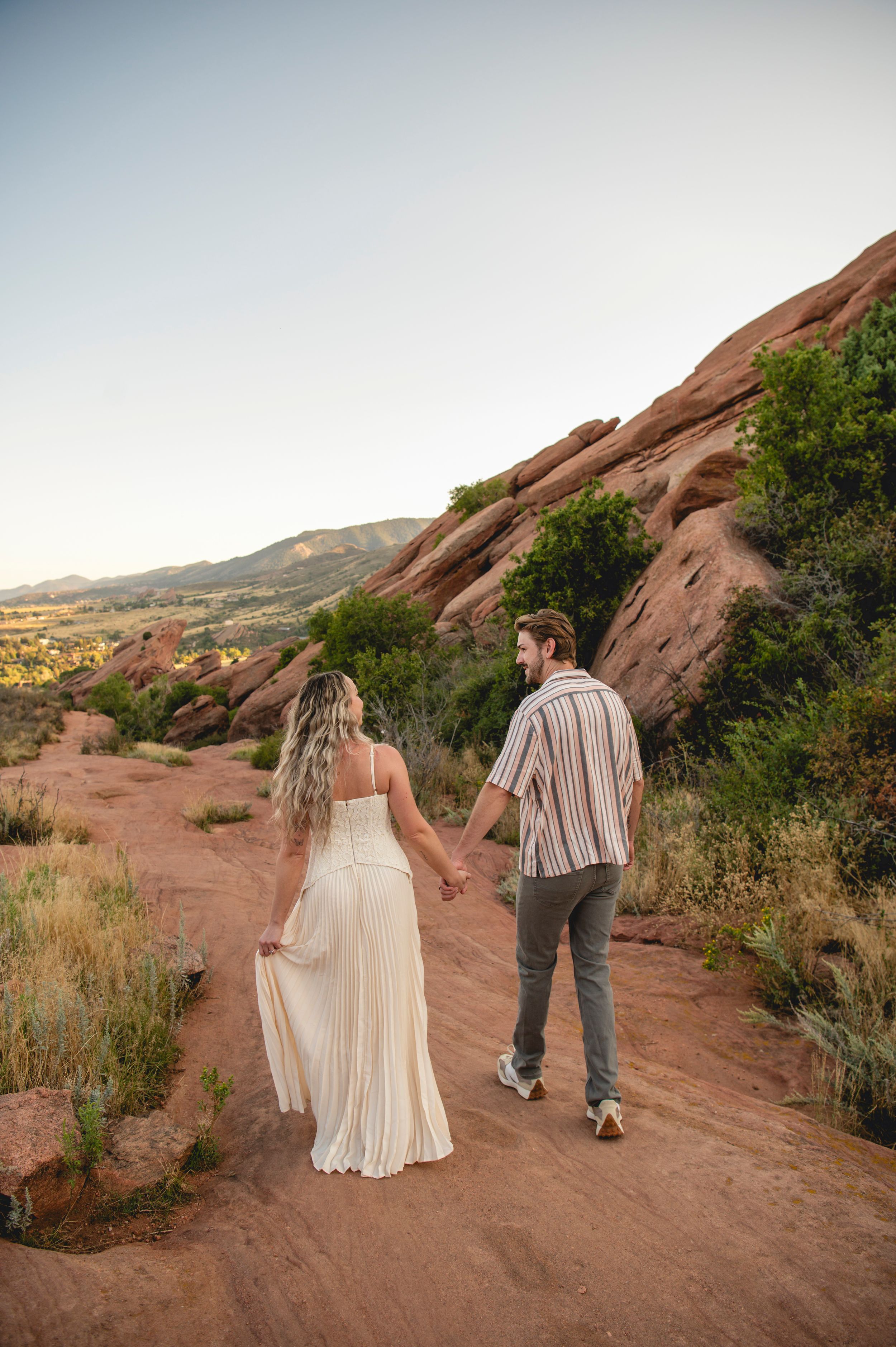 A couple holds hands walking along a red rock trail at sunset in a scenic desert landscape.