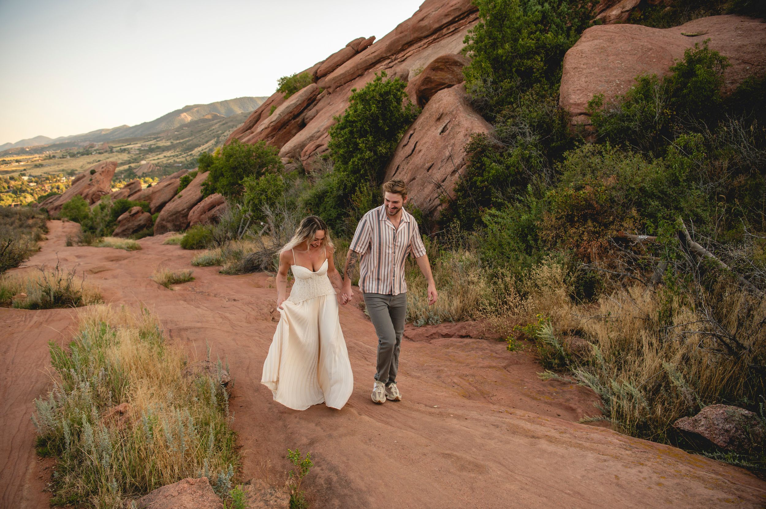 Bride and groom walk hand in hand along a red rock trail at Red Rocks Park, Colorado, at sunset.