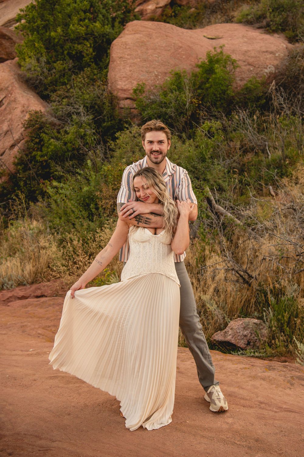 Couple embracing on red rock trail, woman in ivory pleated dress, man in striped shirt, Colorado desert landscape.