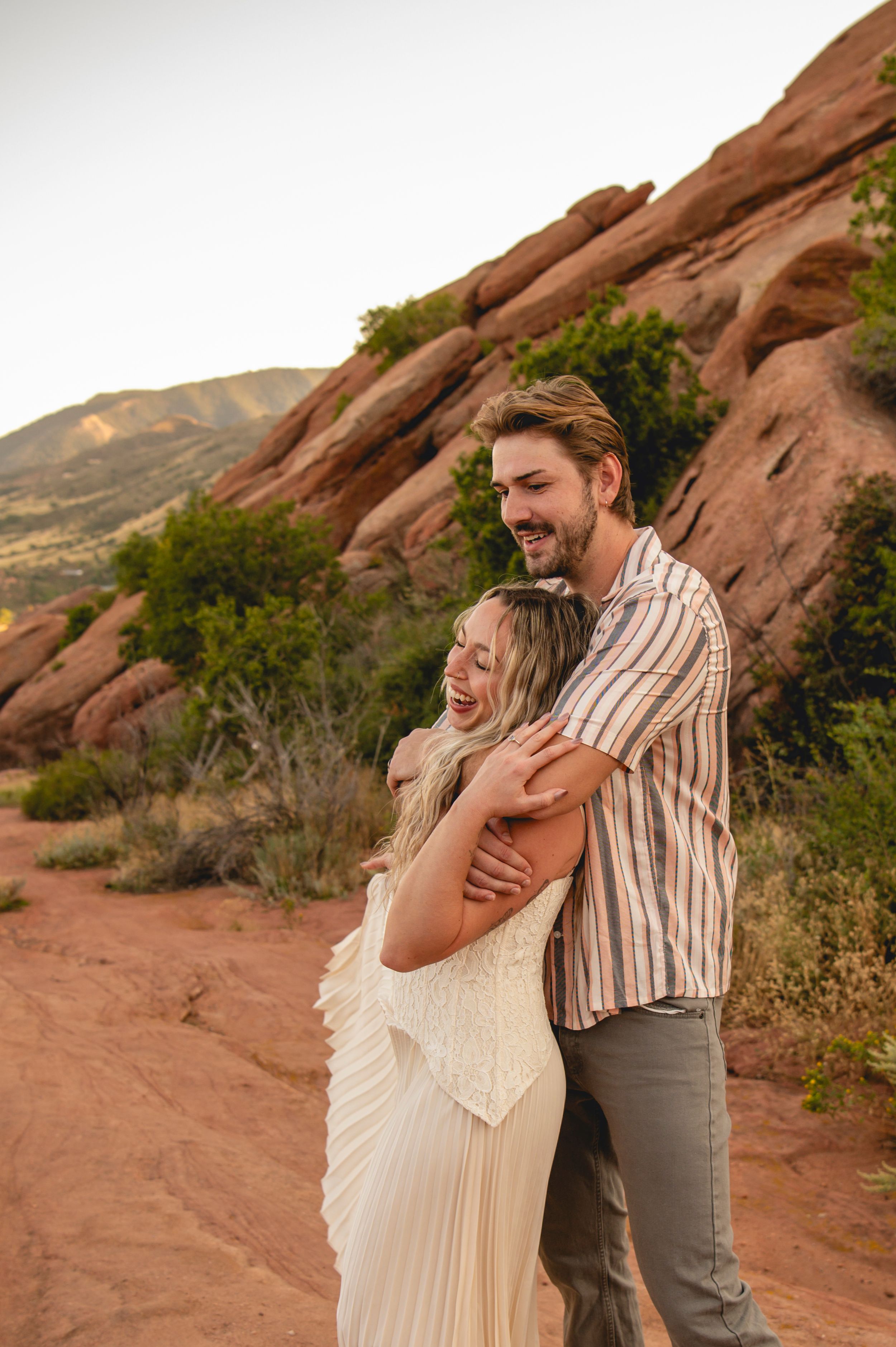 A couple embraces and laughs among red rock formations in a desert landscape, woman in white dress, man in striped shirt.