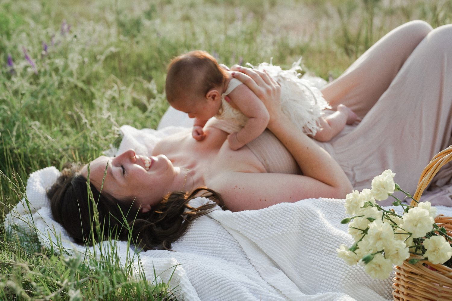 Mother in beige dress holding newborn baby outdoors in a field of wildflowers.
