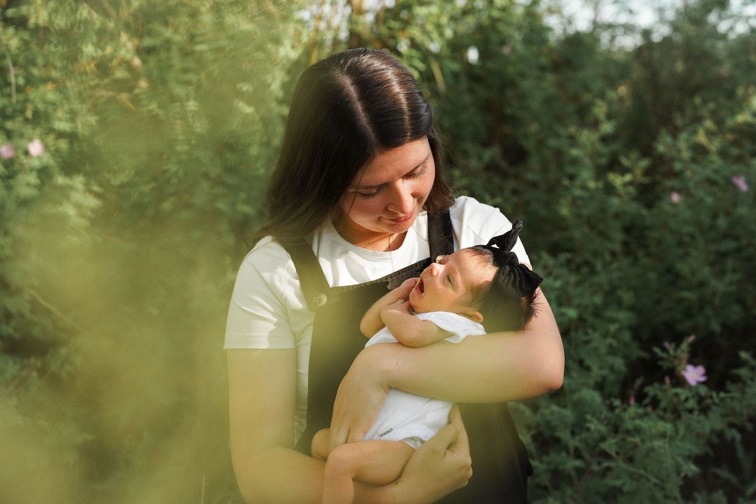 Mother in beige dress holding newborn baby outdoors in a field of wildflowers.