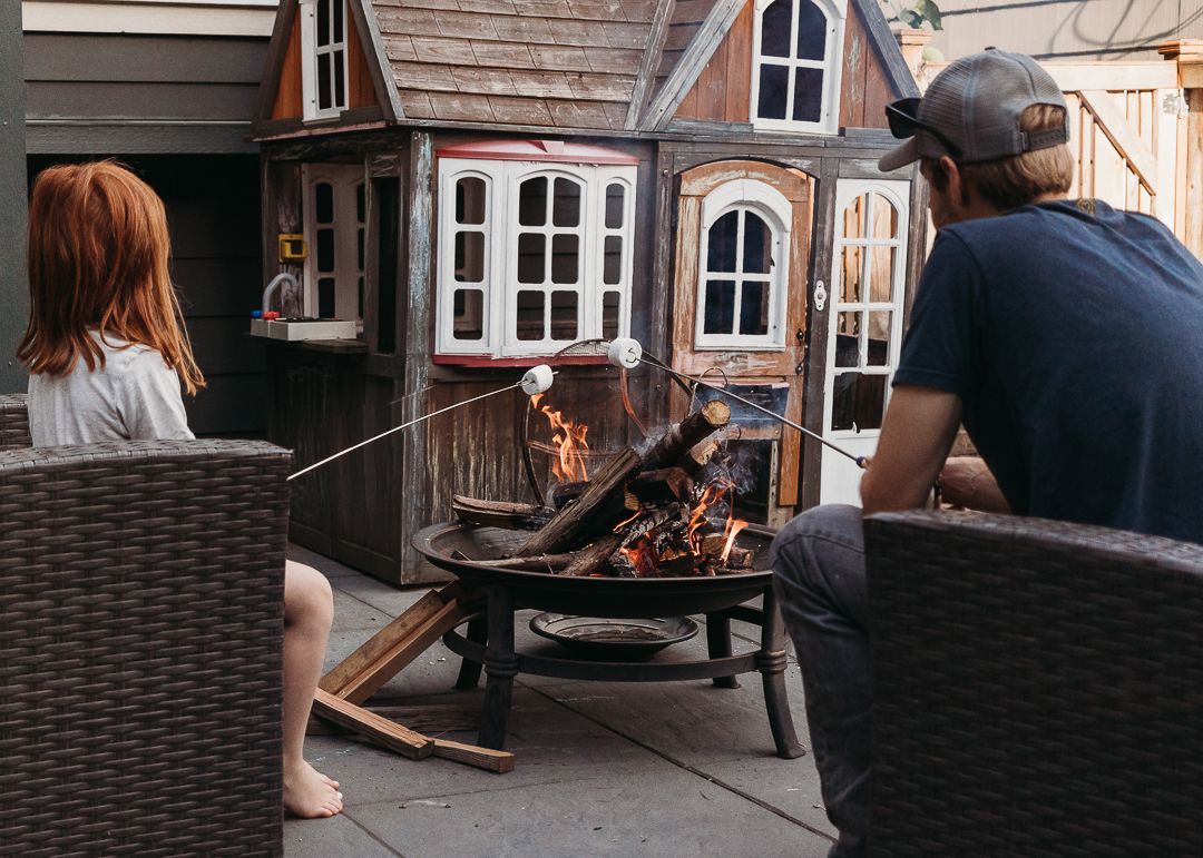 Two people roasting marshmallows over a fire pit beside a charming playhouse on a cozy outdoor patio.