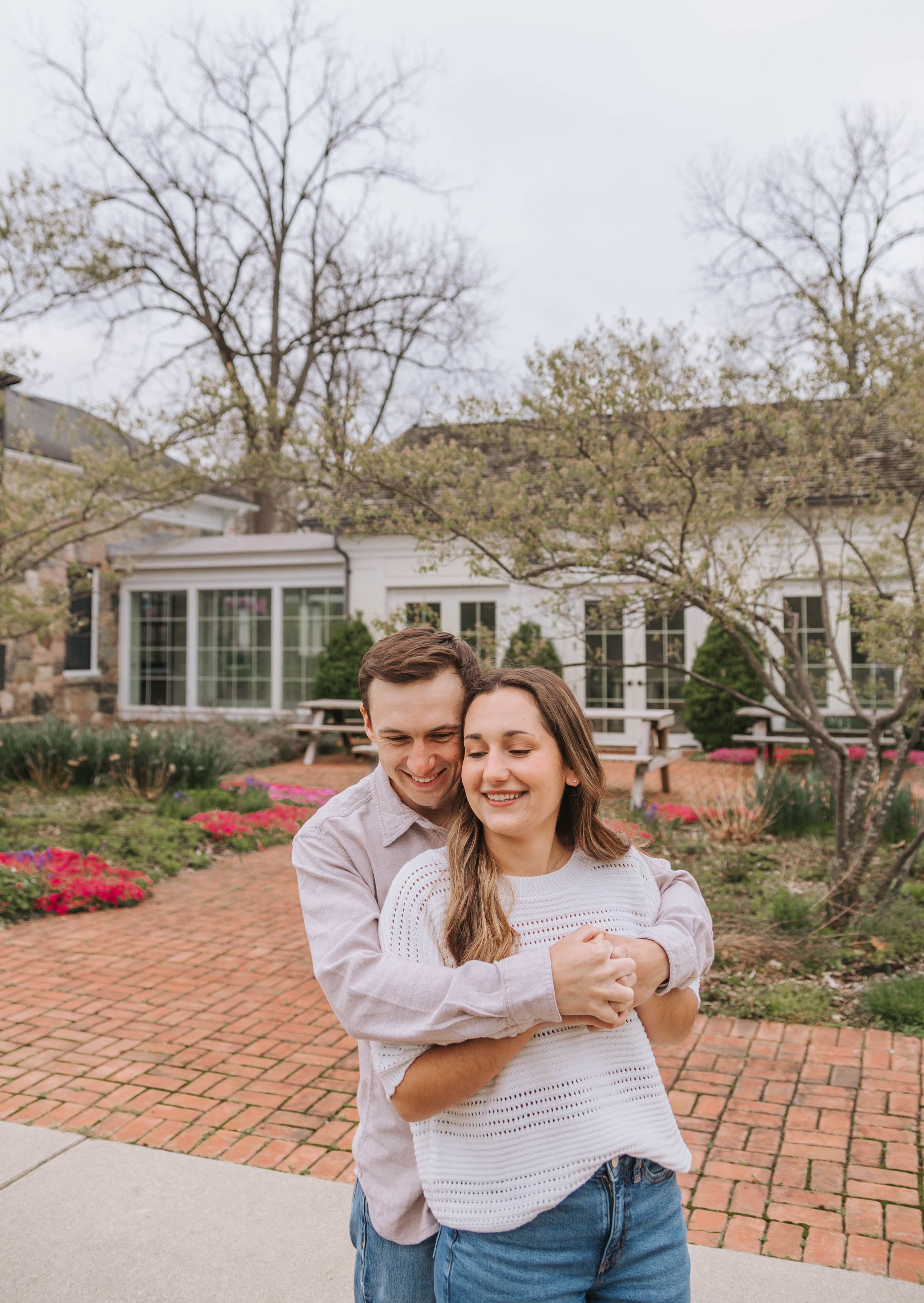 A couple embraces and smiles in a brick garden courtyard with red flowers and a white building backdrop.