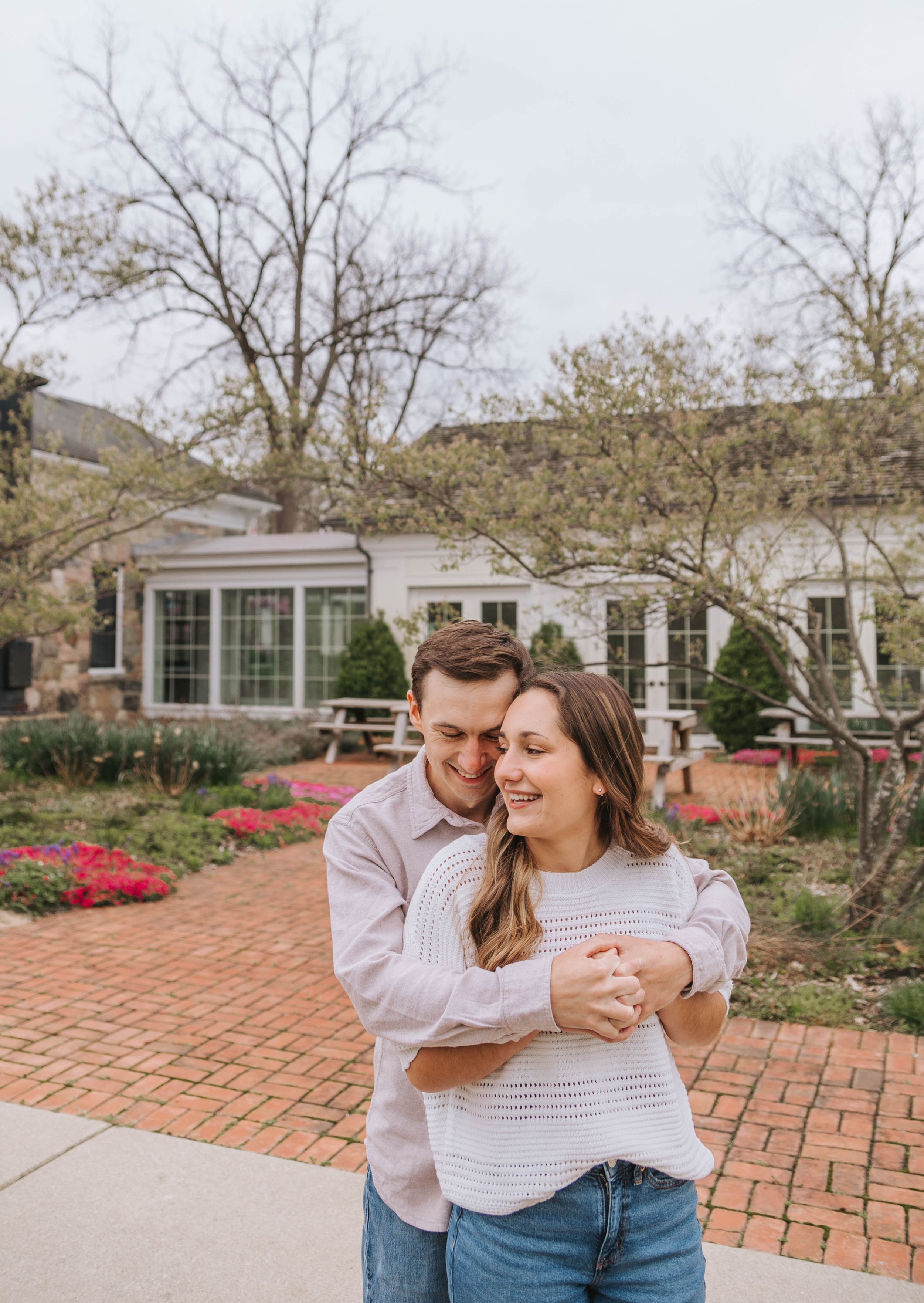 A happy couple hugs and laughs together in a charming brick garden with colorful flowers and white building.