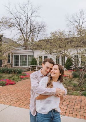 Couple embracing and smiling in a garden courtyard with red flowers and a white building backdrop.
