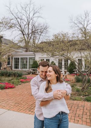 Couple embracing and smiling outdoors near a white building with red brick pathway and spring flowers.