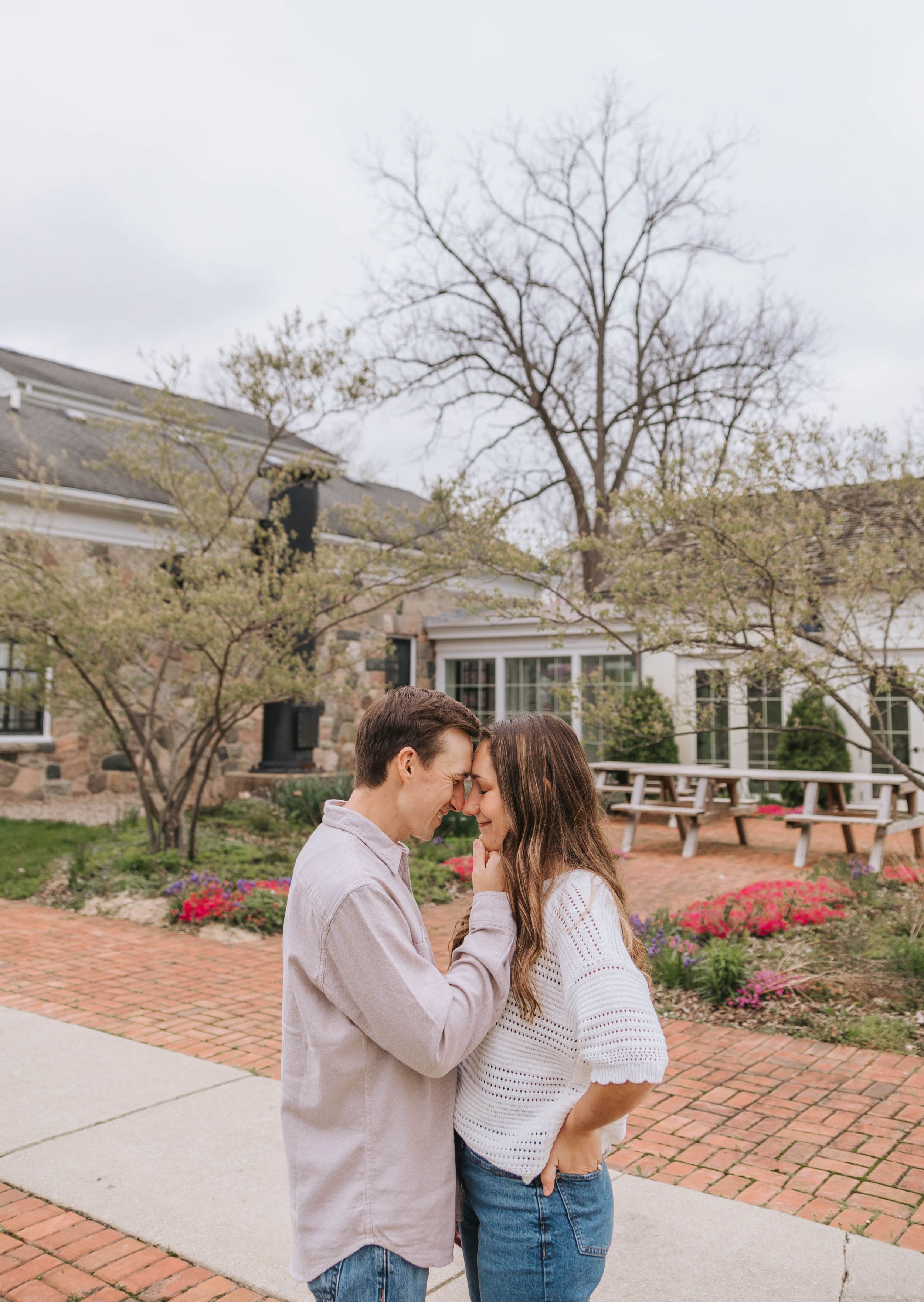 A couple shares a romantic forehead touch outdoors near a stone building with blooming spring gardens.