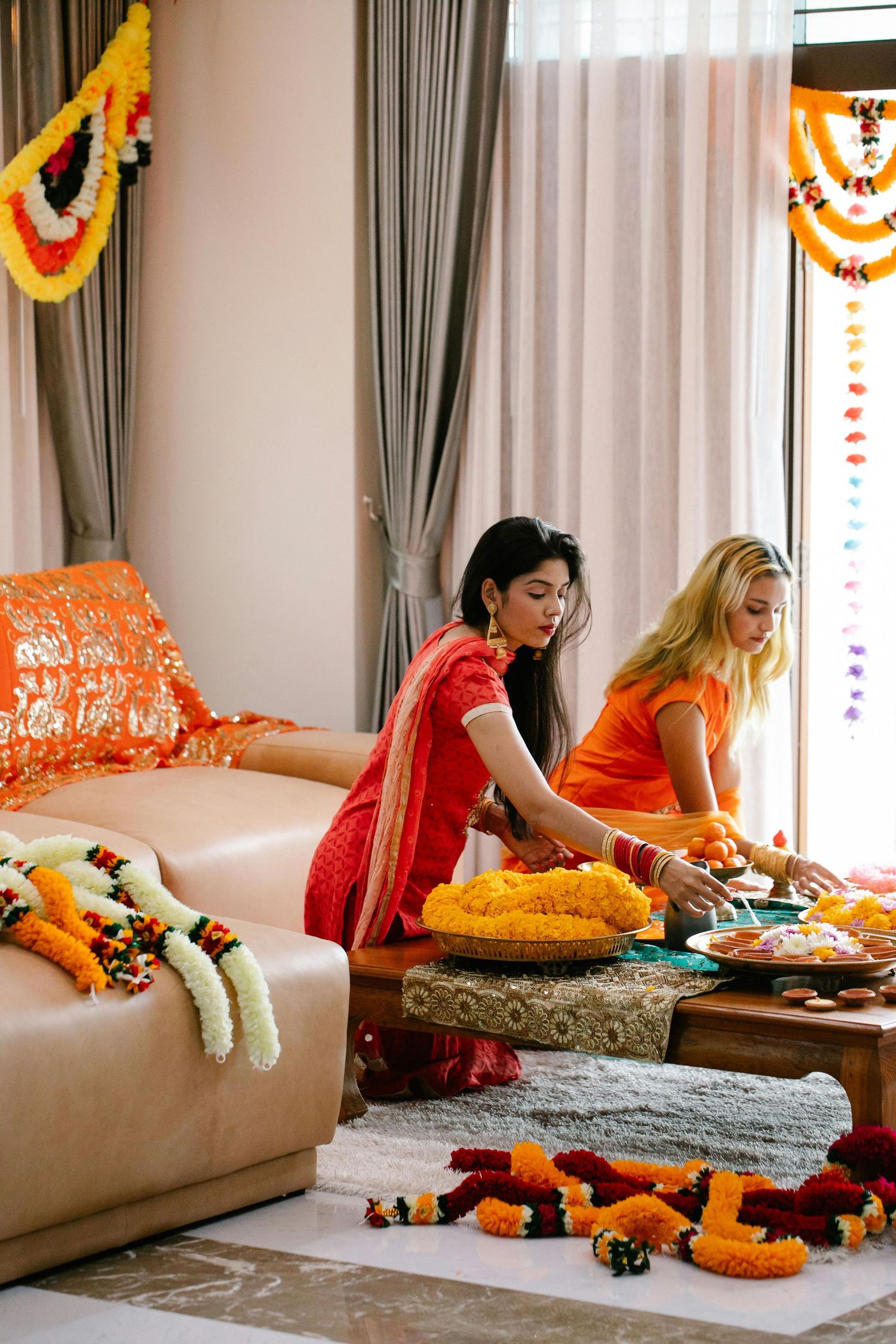 Two women in vibrant traditional attire arrange offerings during a festive Indian celebration indoors.