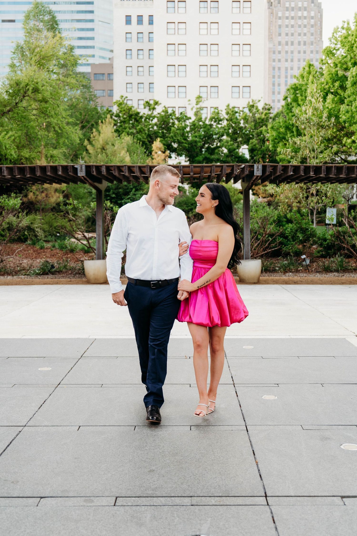 Couple holding hands walking in urban park, woman in pink mini dress, man in white shirt and navy pants.