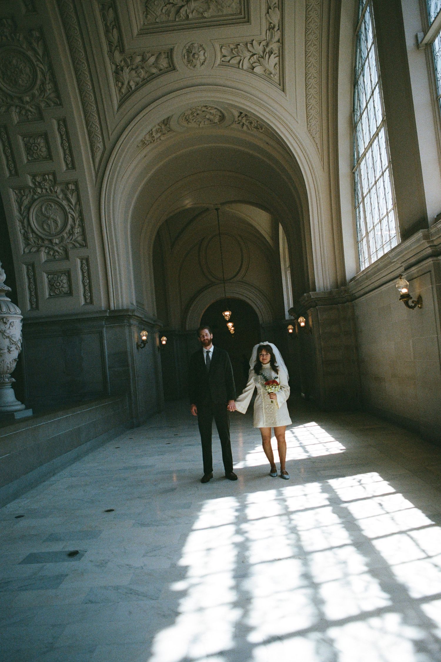 A couple in wedding attire stands in a grand ornate hallway with dramatic natural light streaming through tall windows.