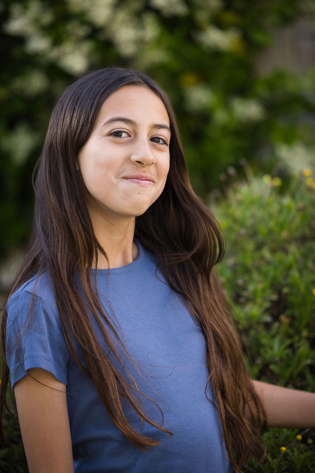 A young woman with long dark hair smiles outdoors, wearing a blue t-shirt with green shrubs behind her.