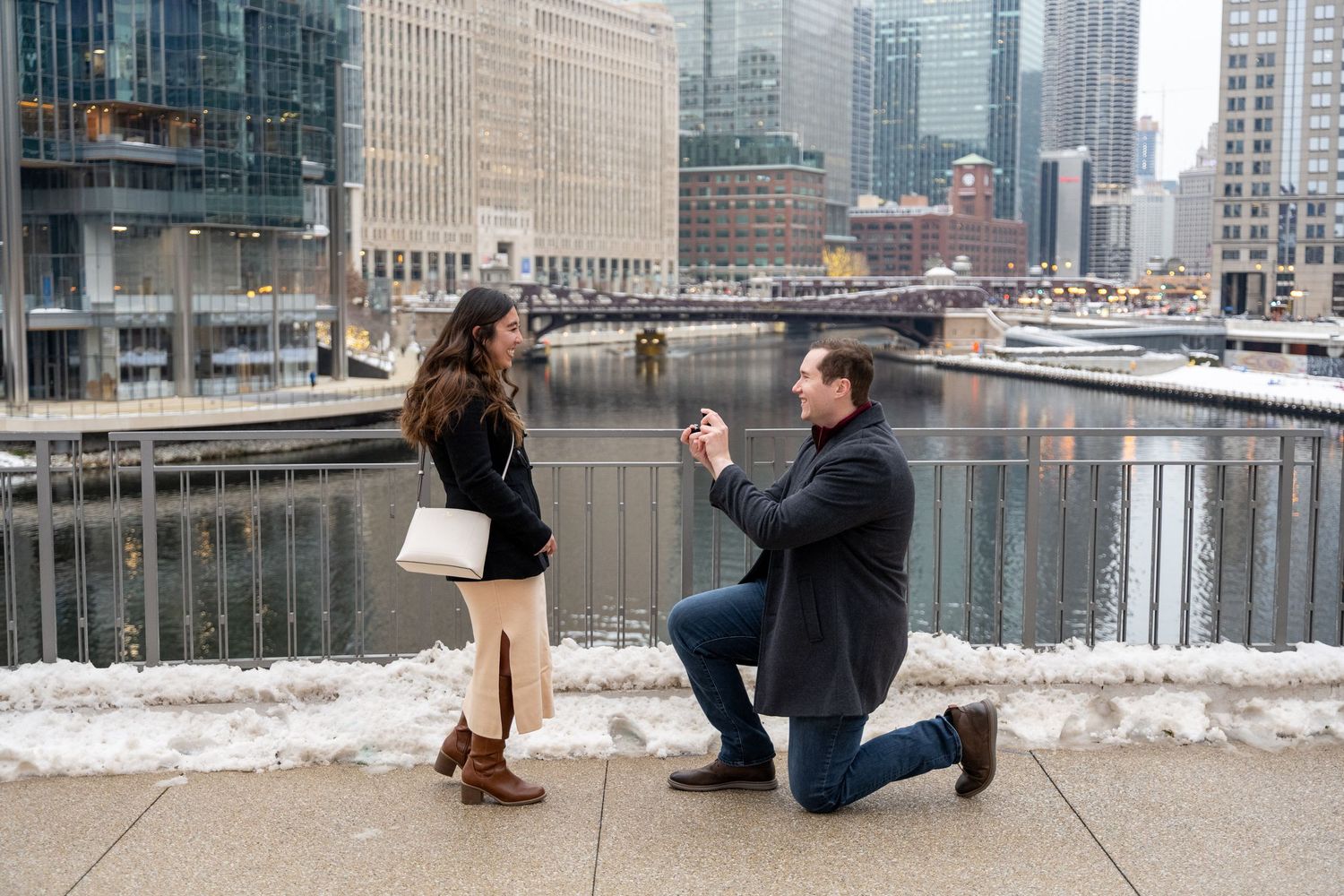 Couple embracing on a bridge over the Chicago River, woman in floral dress and man in gray suit, urban skyline backdrop.