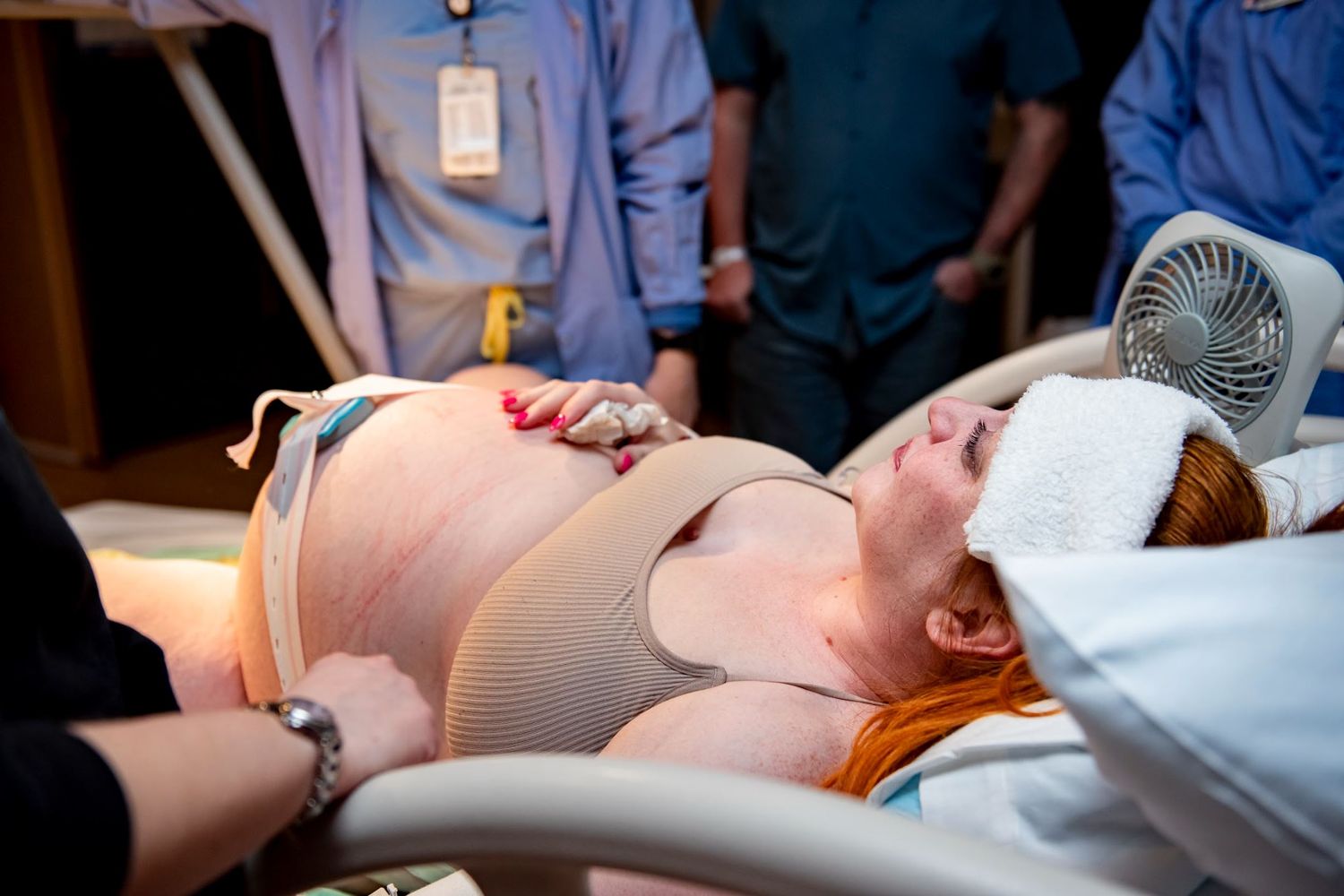 Healthcare team surrounds a patient on a medical bed with monitoring equipment and fan nearby.