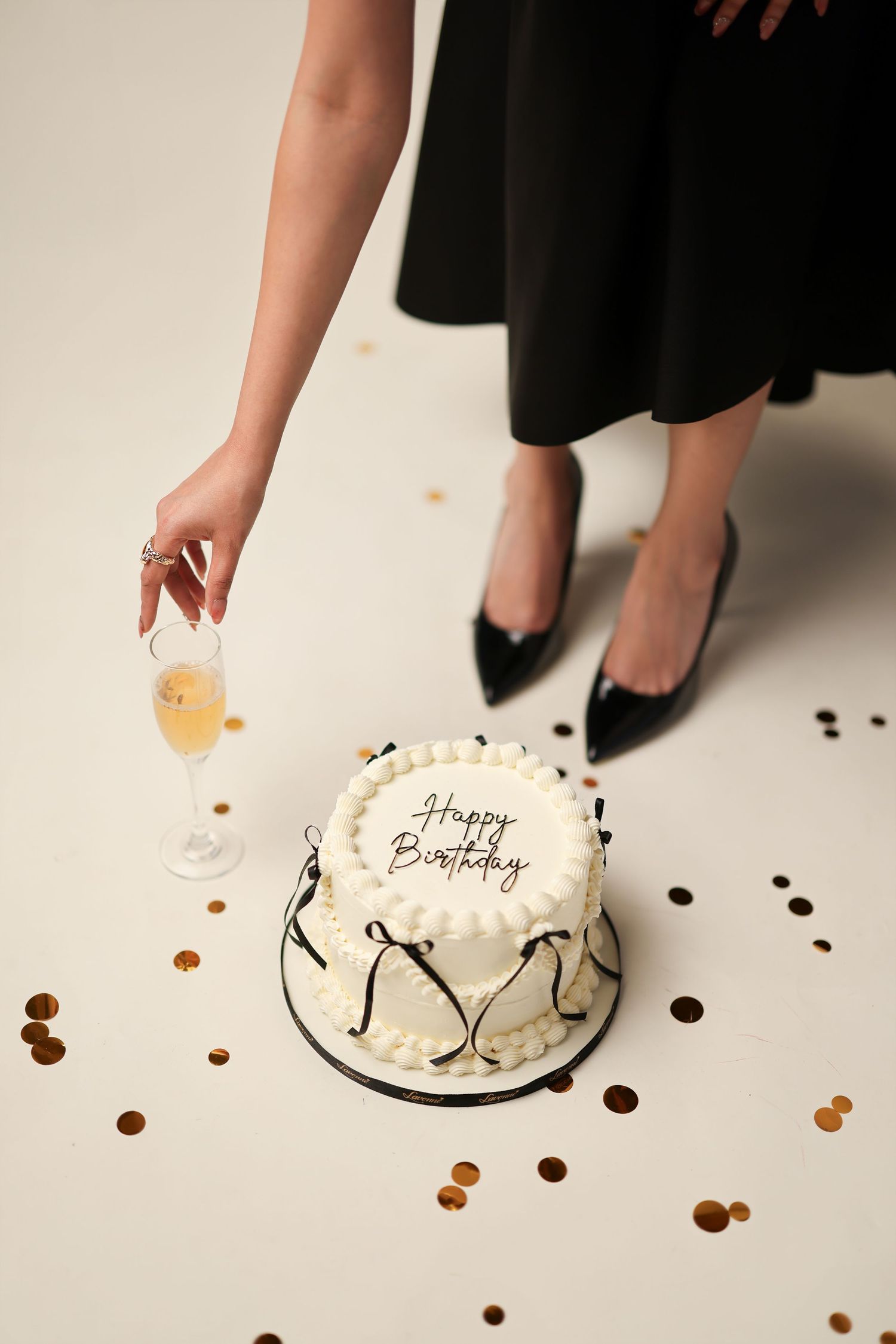 Woman in black off-shoulder gown holds white birthday cake with candle beside black balloons on white background.