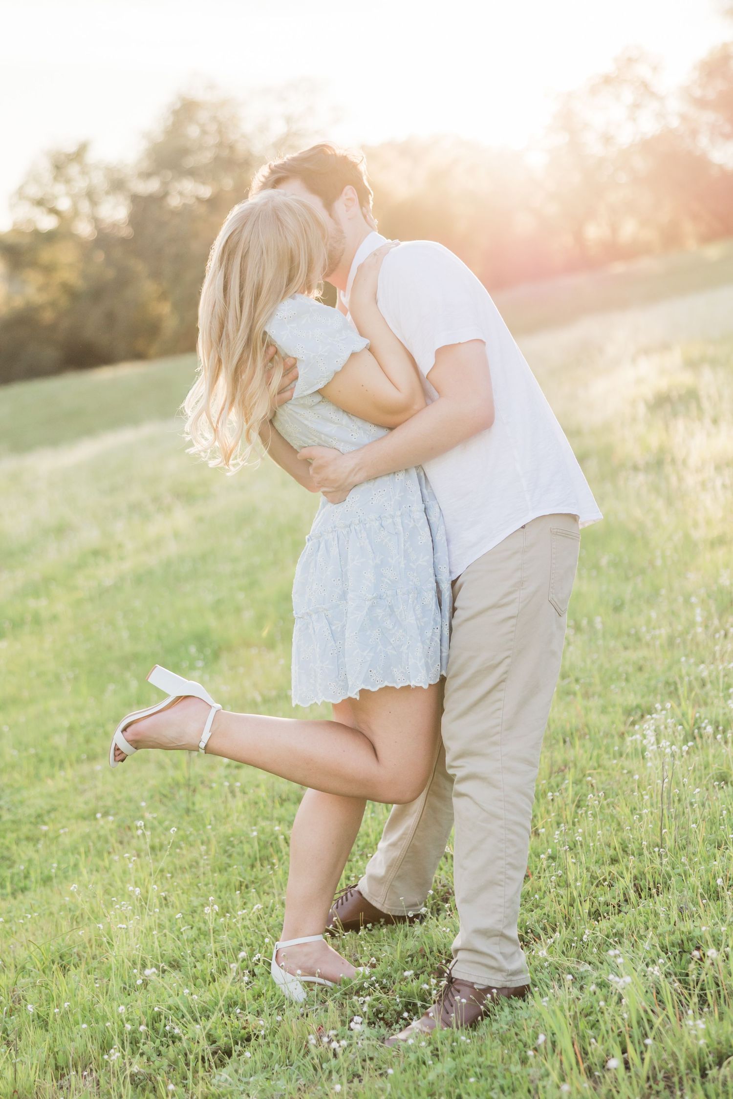 Couple sharing a romantic kiss in a sunlit green meadow, woman in light blue dress lifting her leg.