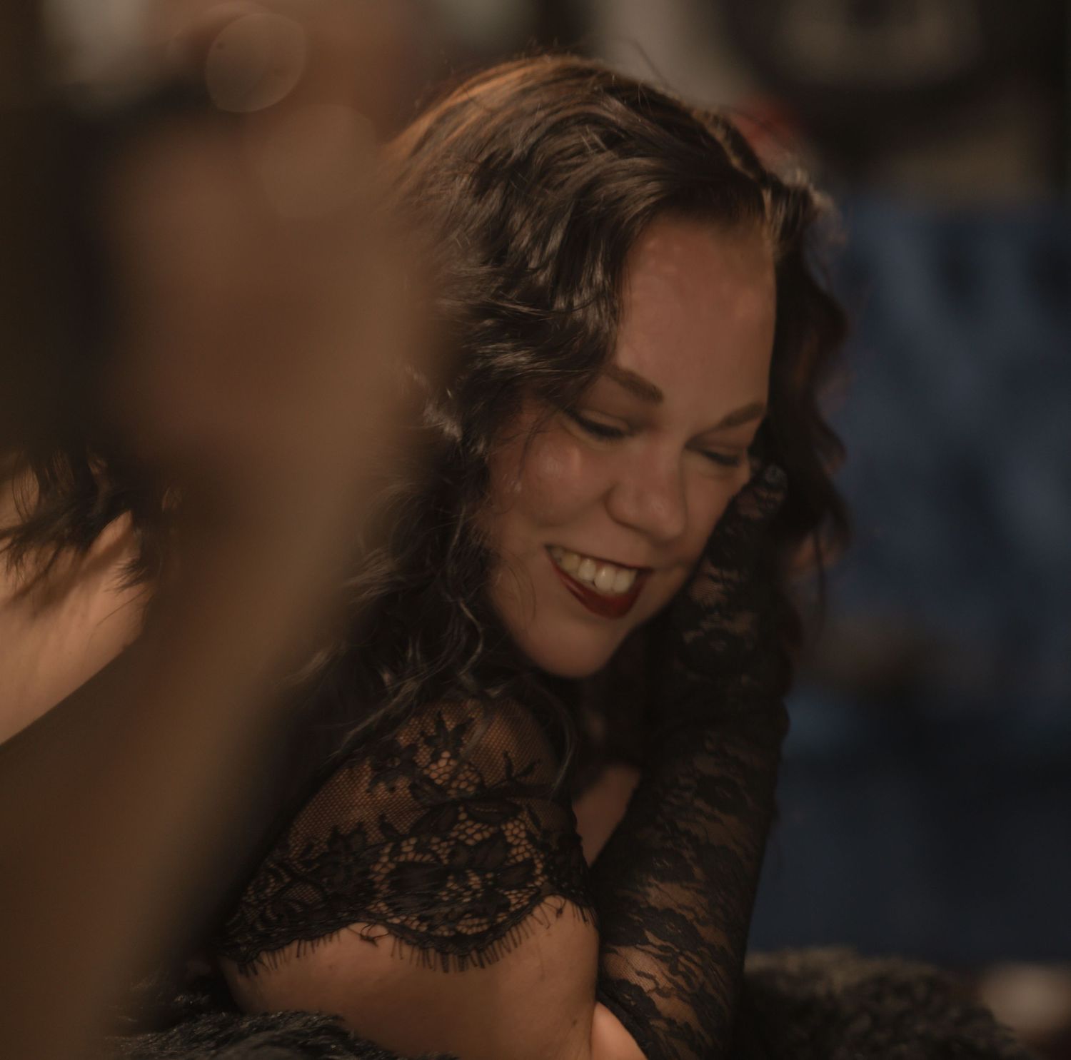 Woman in black lace top smiling joyfully at a dimly lit social event.