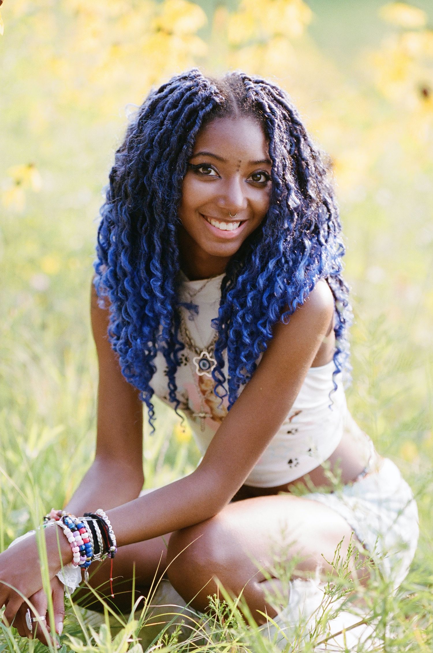 Young woman with blue braided hair smiling outdoors in a sunny field of yellow wildflowers.
