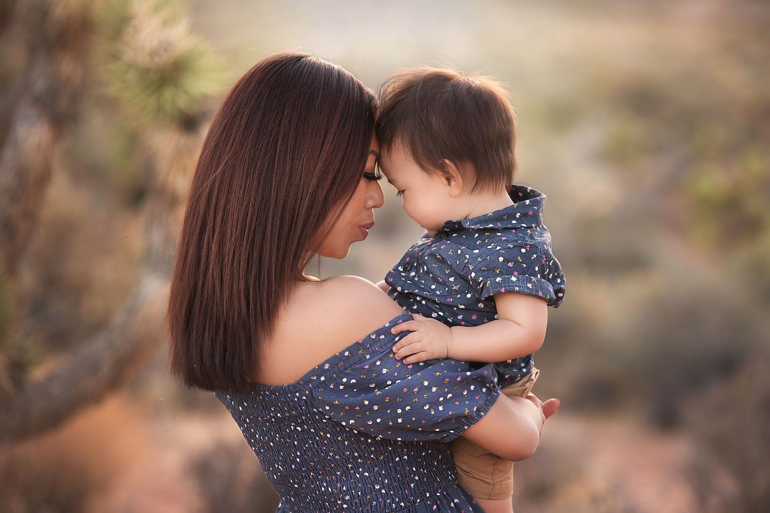A mother in a polka dot dress tenderly holds her baby outdoors in a soft, natural desert setting.