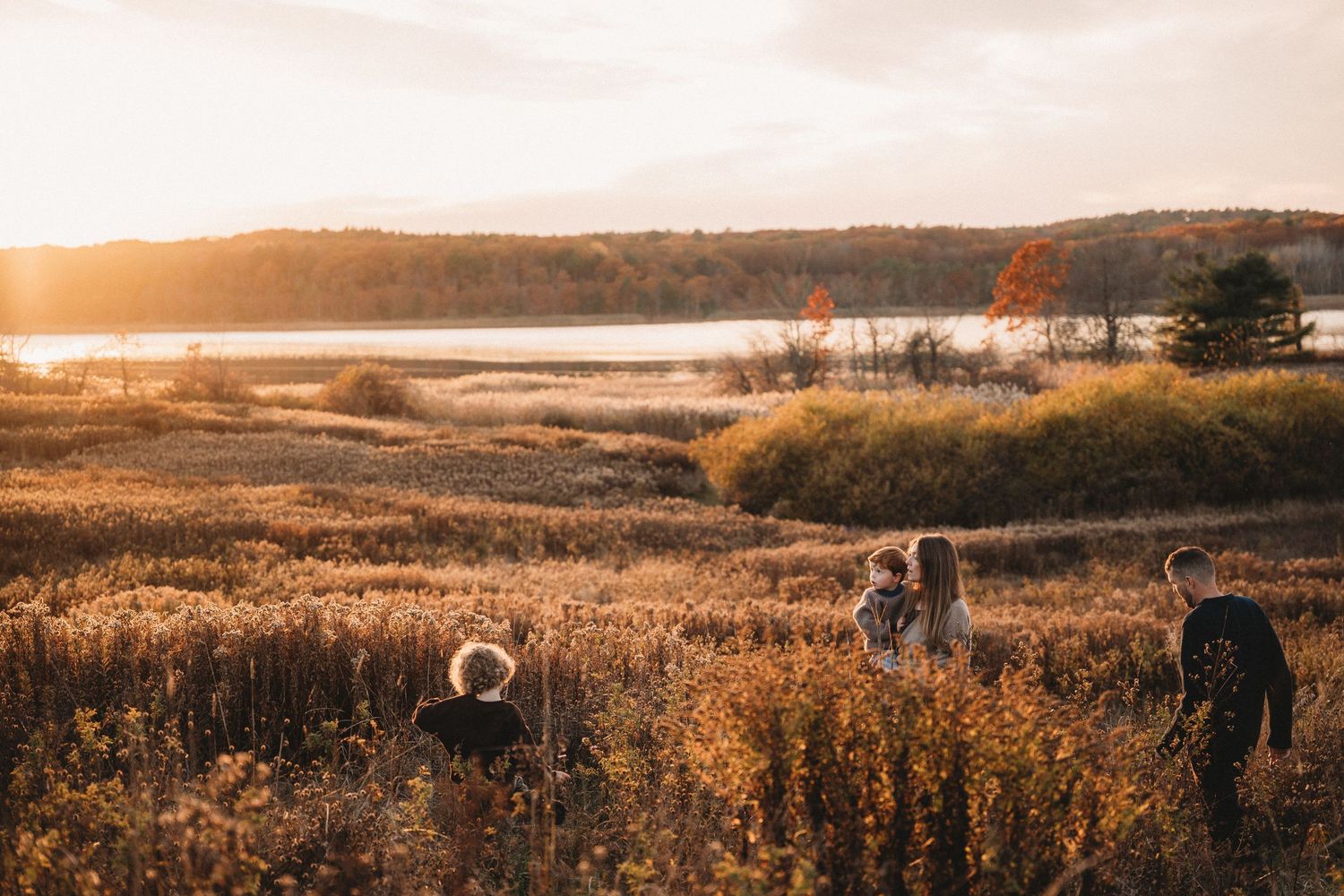 Pregnant couple walking hand-in-hand on a rocky beach at sunset, wearing casual outfits with golden light behind them.