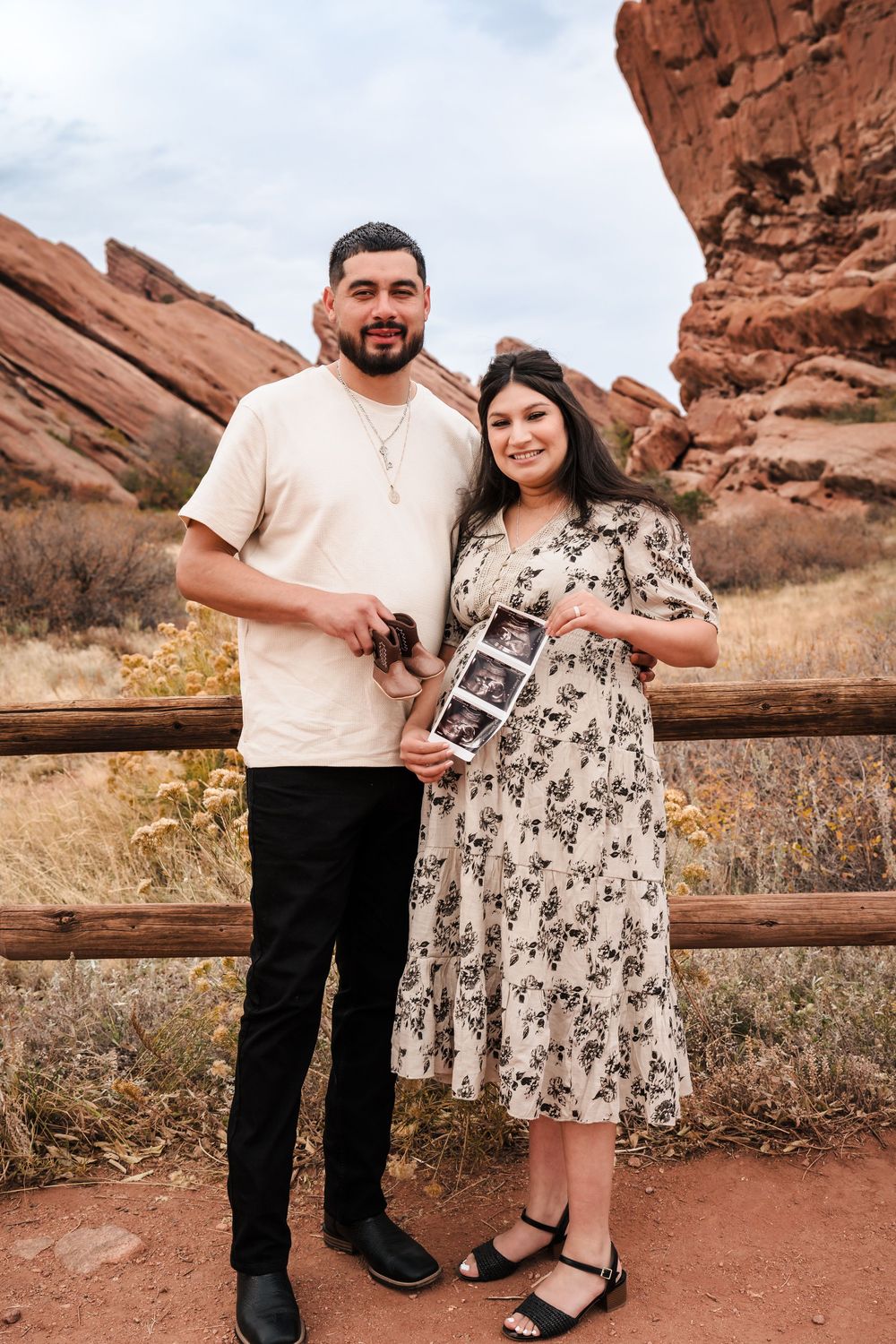 Couple holding ultrasound photos at Red Rocks Park, announcing pregnancy amid iconic red rock formations in Colorado.