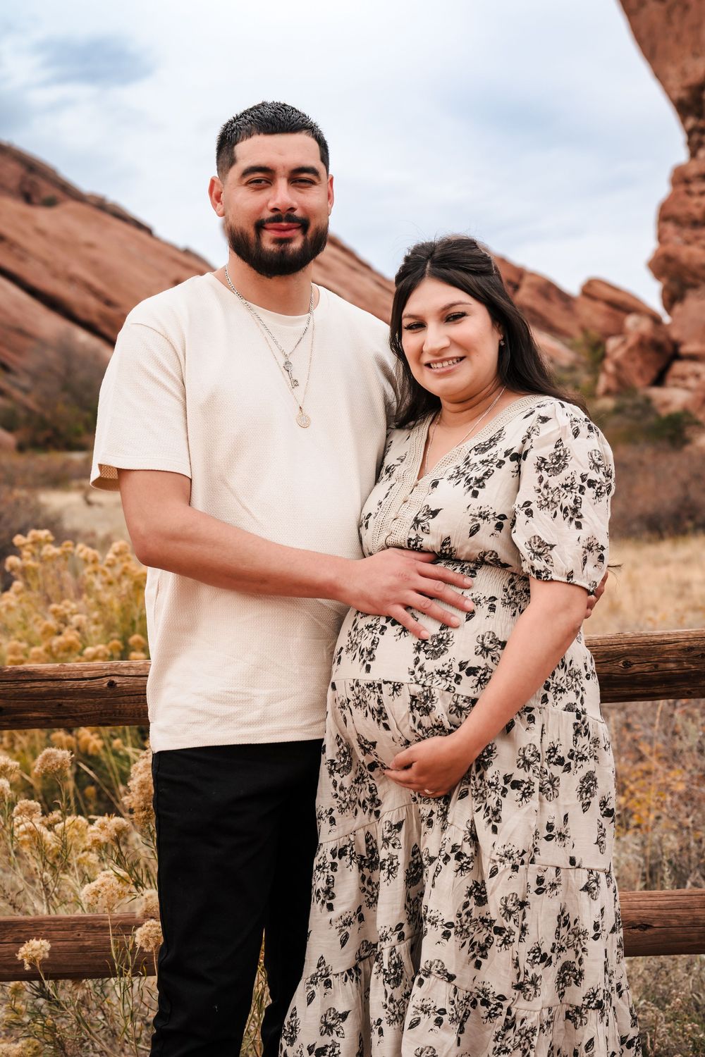 Couple poses for maternity photo at Red Rocks Park, woman in floral dress cradling baby bump.