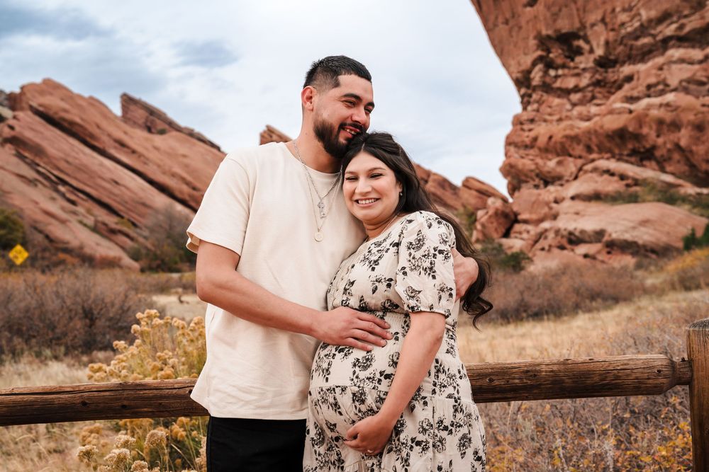 Couple poses at Red Rocks Park, pregnant woman in floral dress with partner in cream shirt, red rock formations behind them.