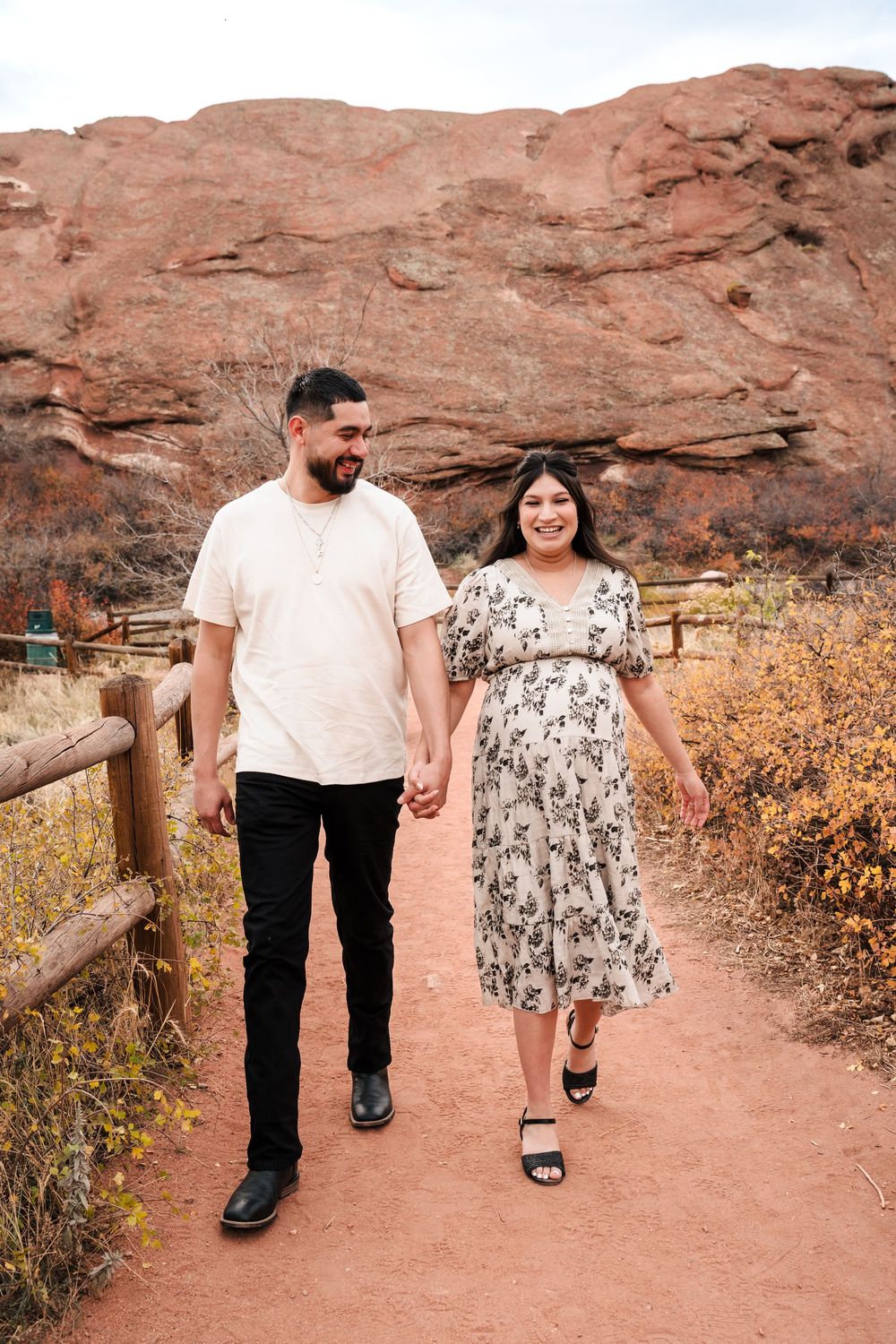 Couple holding hands walking on a red dirt path at Red Rocks Park, woman in floral dress, man in white shirt and black pants.