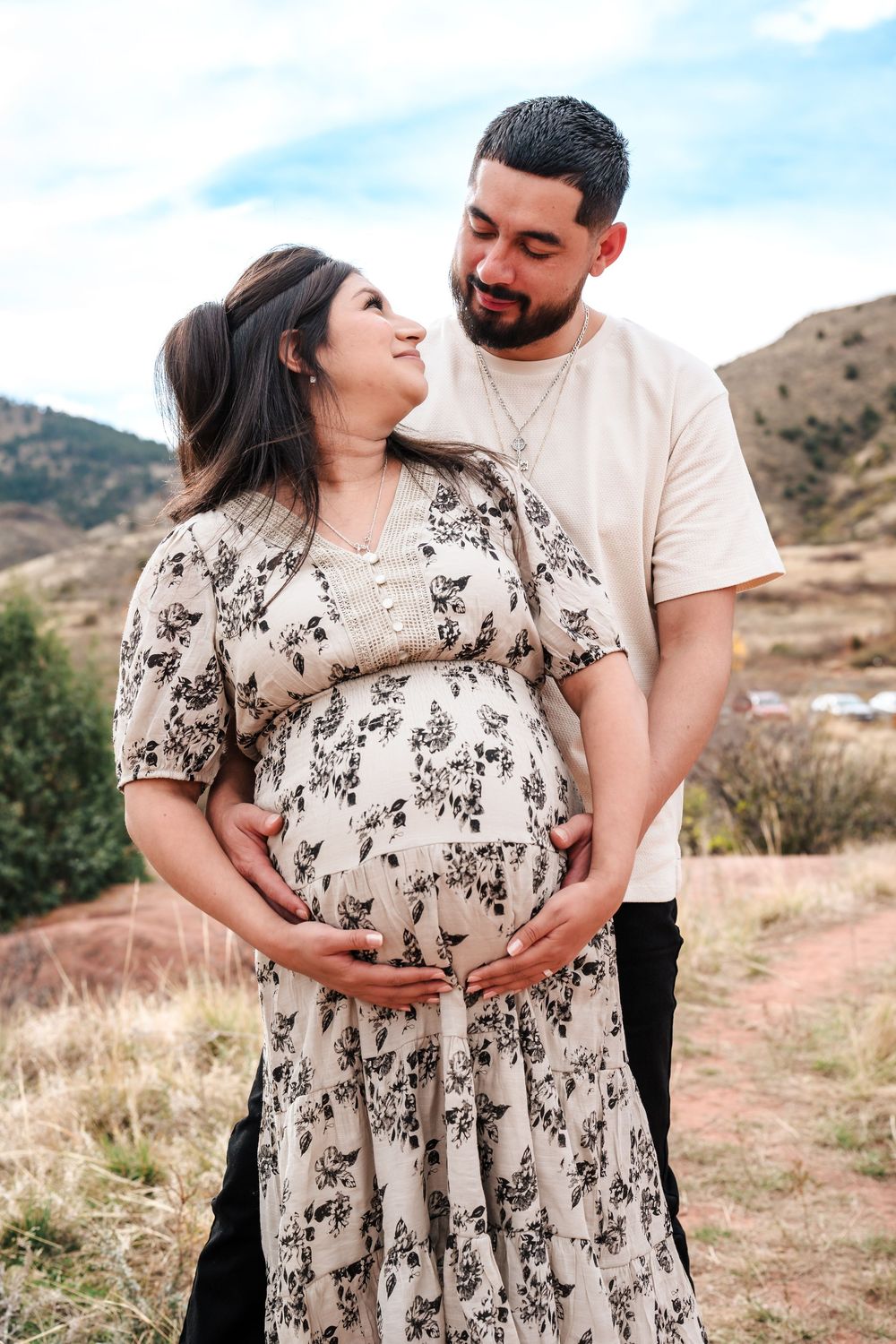 A couple poses outdoors, man embracing pregnant woman in floral dress, both smiling with mountain backdrop.