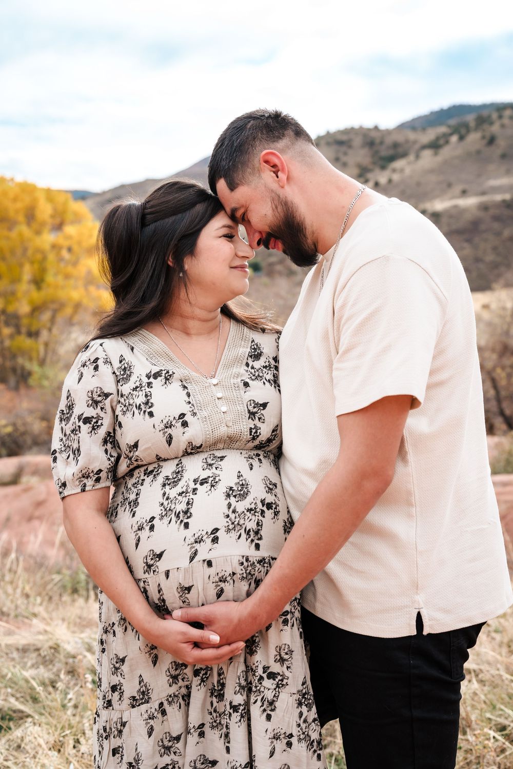 Couple sharing a tender moment outdoors, pregnant woman in floral dress with partner touching her baby bump, mountain backdrop.