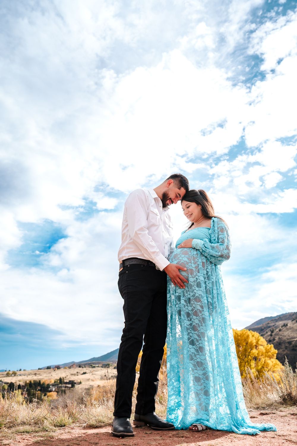Expectant couple shares tender moment outdoors, woman in turquoise lace maternity gown under blue sky.