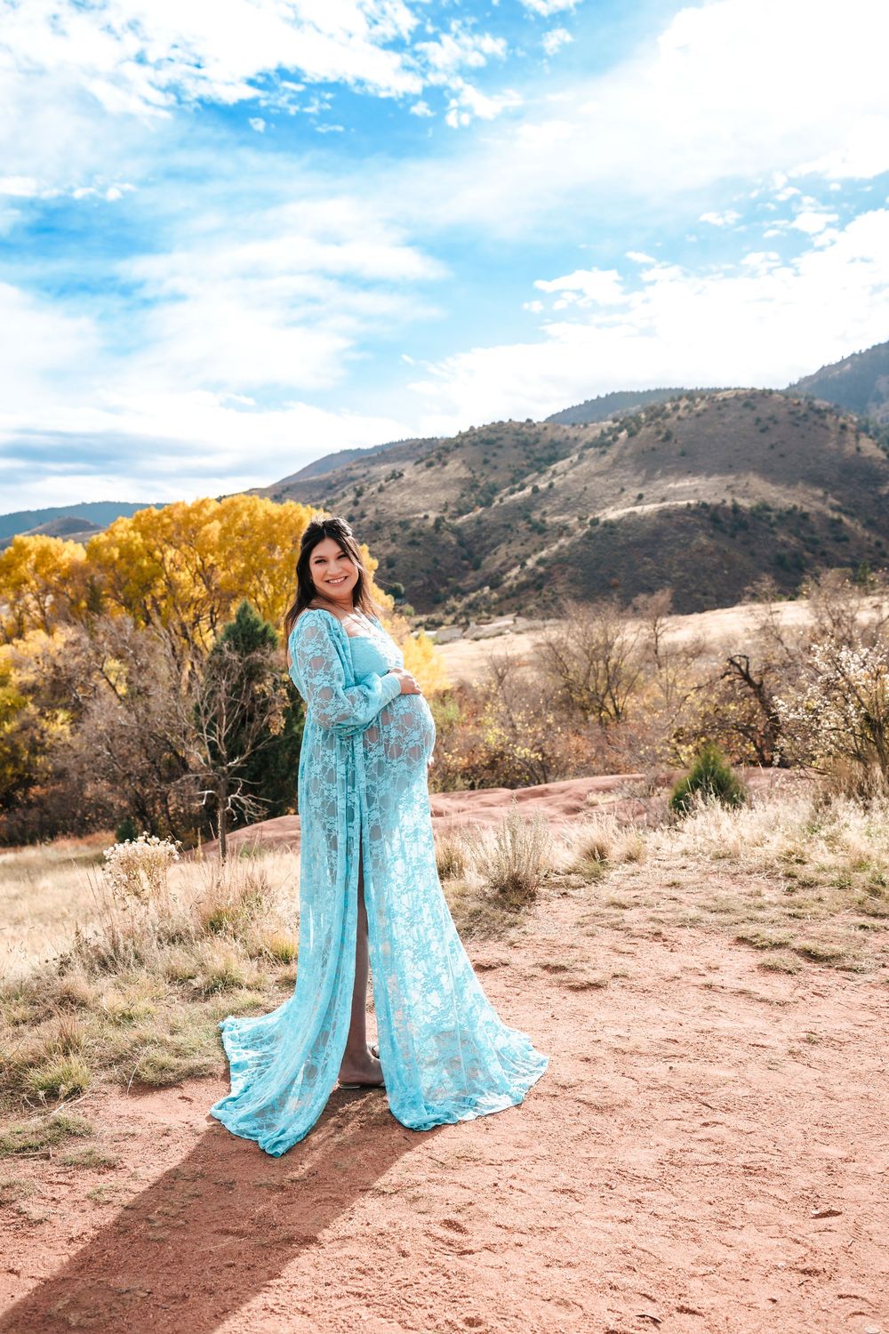 Pregnant woman in teal lace maternity gown poses on red dirt path with mountain backdrop.