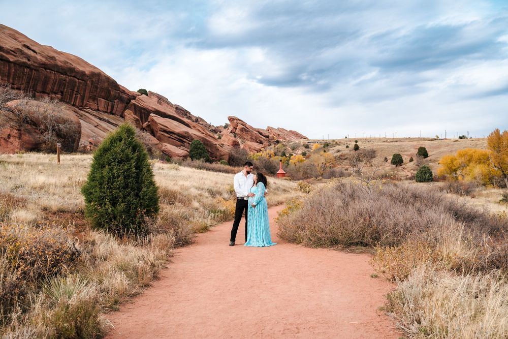 Pregnant couple embraces on red dirt path at Red Rocks Park in Colorado autumn landscape.
