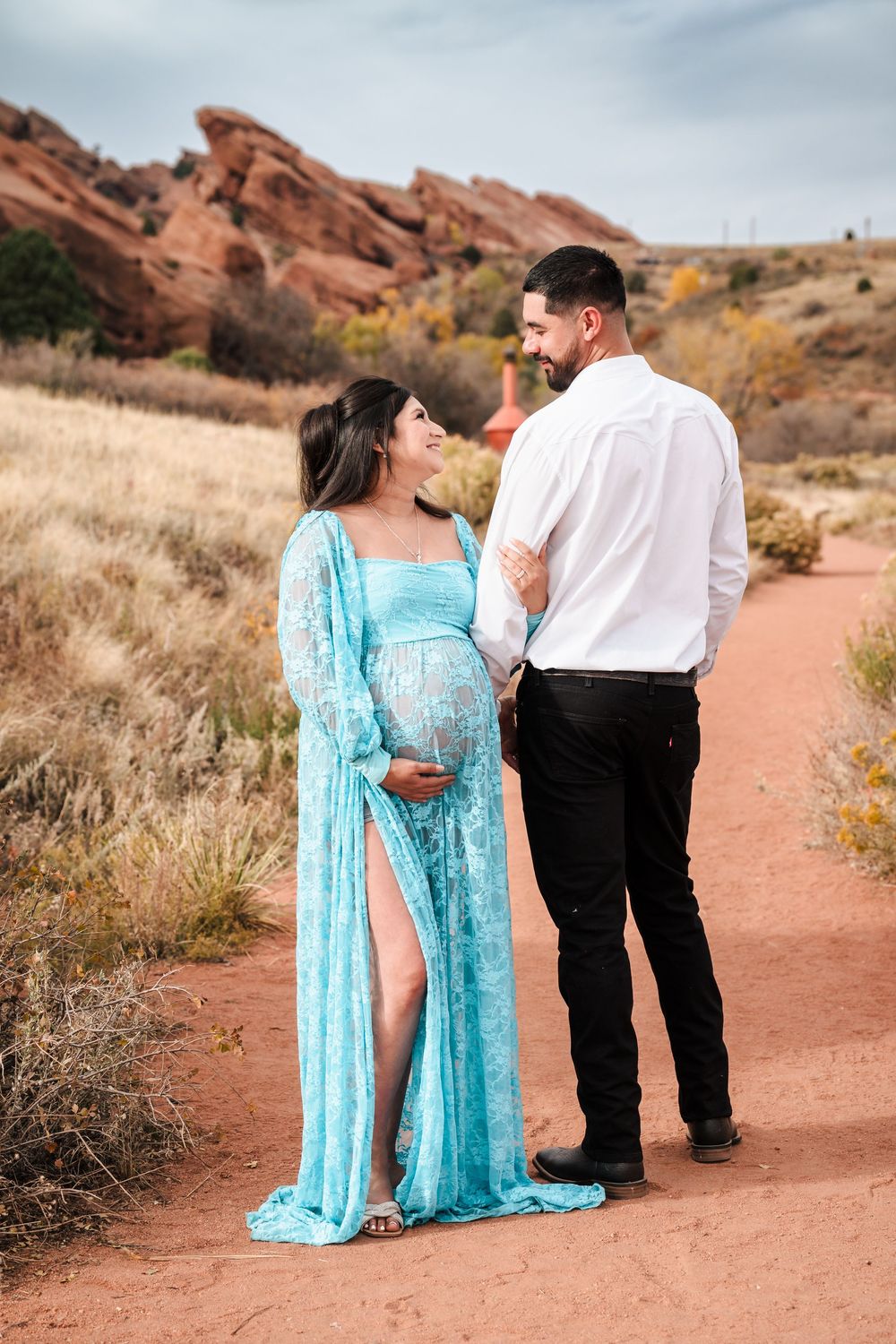 Smiling pregnant couple in turquoise gown and white shirt pose on red dirt trail at Red Rocks.