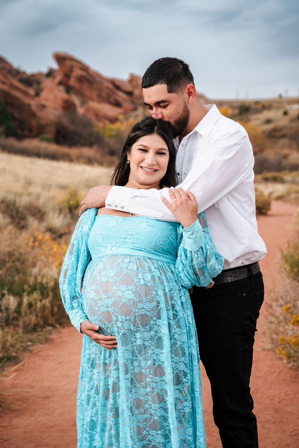Couple shares tender maternity moment on red rock trail, woman in teal lace dress.