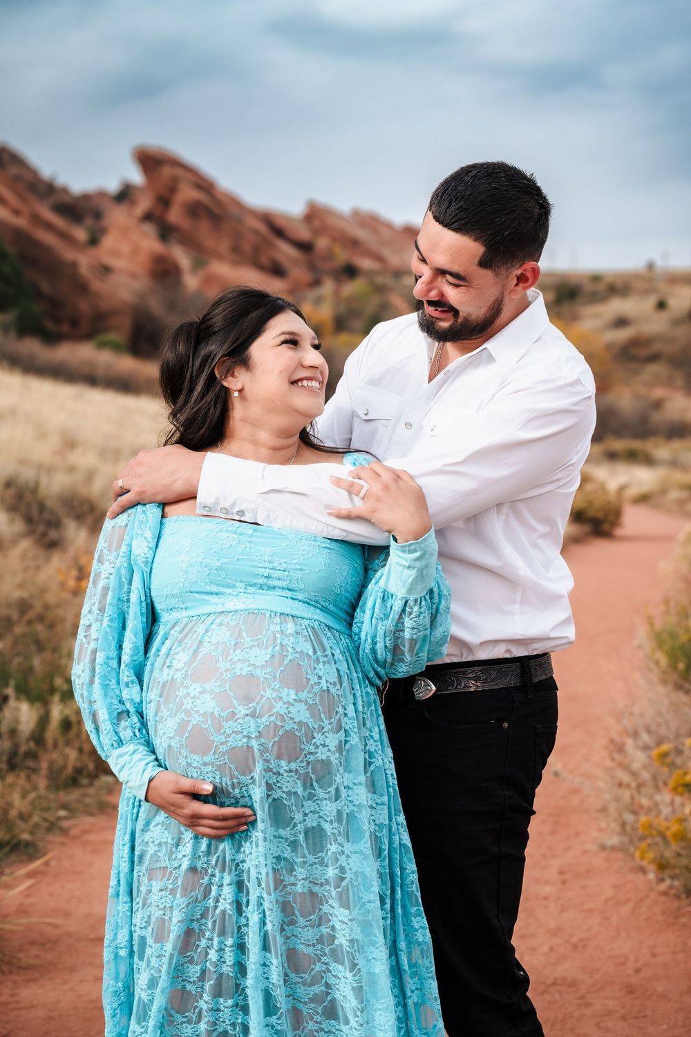Smiling pregnant couple in teal lace dress and white shirt pose at Red Rocks park.